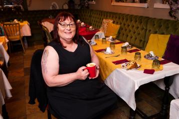 A trans woman sitting down at a table that has a yellow tablecloth. She is holding a red mug and smiling at the camera
