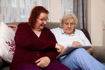 Two older women sitting on a sofa, one wearing a maroon cardigan and pointing at something in a newspaper