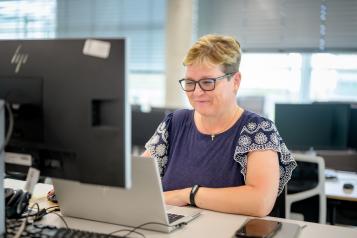A woman with short blonde hair and glasses is sitting in front of a computer, she is slightly smiling. 