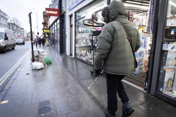 A person wearing a coat with their hood up and walking along a busy street lined with shops. It looks cold and its raining.