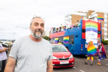 A man wearing a grey t shirt standing in front of a bus with rainbow flags