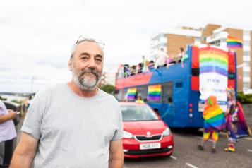 A man wearing a grey t shirt standing in front of a bus with rainbow flags 