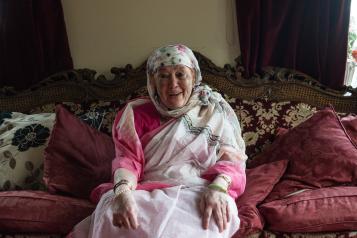 An older woman wearing a white patterned headscarf and a pink sari sitting on a sofa smiling at the camera