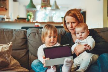 A woman sits on a sofa with a toddler and a baby on her lap. She is looking at a laptop computer. 
