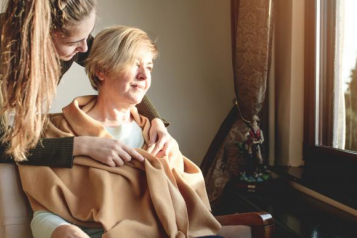 A woman is wrapped in a blanket and sits by a window with her carer 