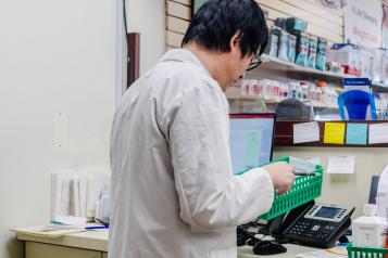 A male pharmacist stands in front of a desk holding a green meds basket 