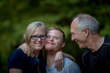 A young boy smiles at the camera and stands in between a woman and man, The woman is smiling at the camera with her arm around the boy. 