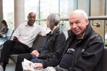 Three people sit in a waiting room. One man is holding a newspaper and smiling. 