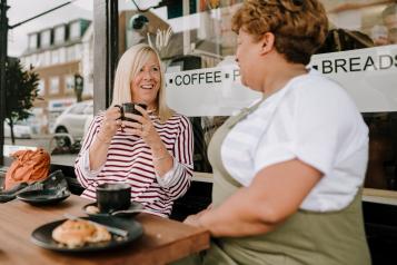 Two middle-aged women sit outside in a cafe drinking coffee, one is smiling towards the camera