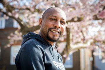 A man smiles at the camera whilst sitting outside. A blossom tree is in the background 