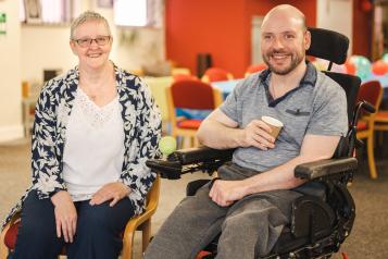 A woman sits next to a man in a wheelchair. He is holding a cup and smiling at the camera.