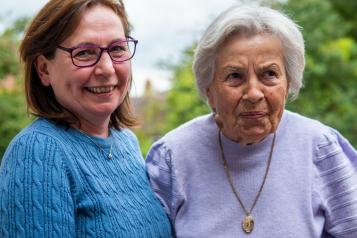 A woman with brown hair is holding the hand of an elderly lady. The woman on the left is wearing a blue jumper and smiling, the elderly lady is wearing purple and is also smiling. at the camera.