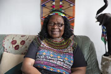 A black woman sitting on a sofa smiling, wearing colourful clothing and a head band,