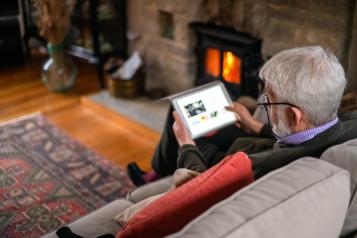 An older man wearing glasses sitting in front of a fire in a cosy-looking room, holding a laptop