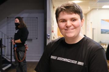 A woman who is blind wearing a black t shirt smiles at the camera. She is at a climbing club. 