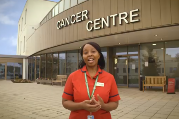 A woman wearing an orange top standing outside a building with a sign above her that says 'Cancer Centre' 