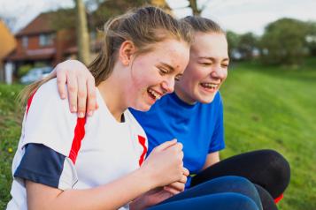 Two teenage girls sit on the grass laughing 