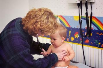 A woman is holding a baby who is sitting up in a nappy, the women is wearing a stethoscope. A rainbow print is in the background.