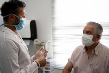 Male patient sitting on a bed talking to a male doctor. Both are wearing masks.