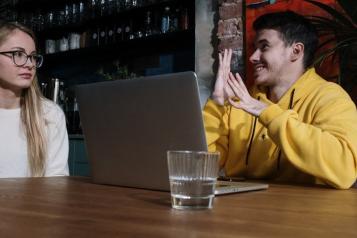 A woman on the left and a may on the right who is signing. He is wearing a yellow hoodie, sitting in front of a laptop. 