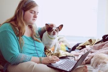 A woman wearing a turquoise top sitting down with her laptop. She is wearing headphones. A white and brown dog is sitting next to her. 