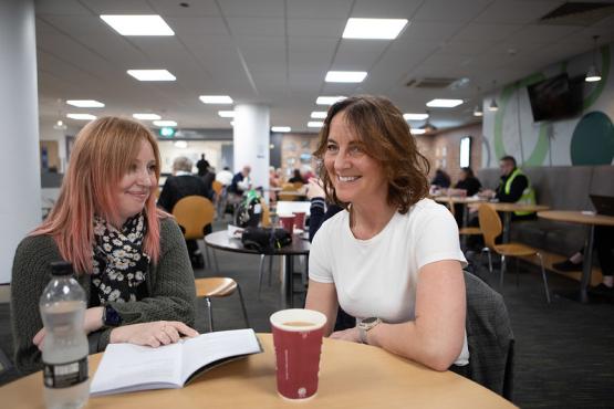 Two women sit down at a table with a bottle of water and coffee cup in the foreground. They are both smiling. 