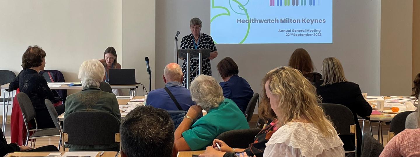 A room of people sitting down with our Chairperson talking in front of a projector with our Healthwatch logo on it 