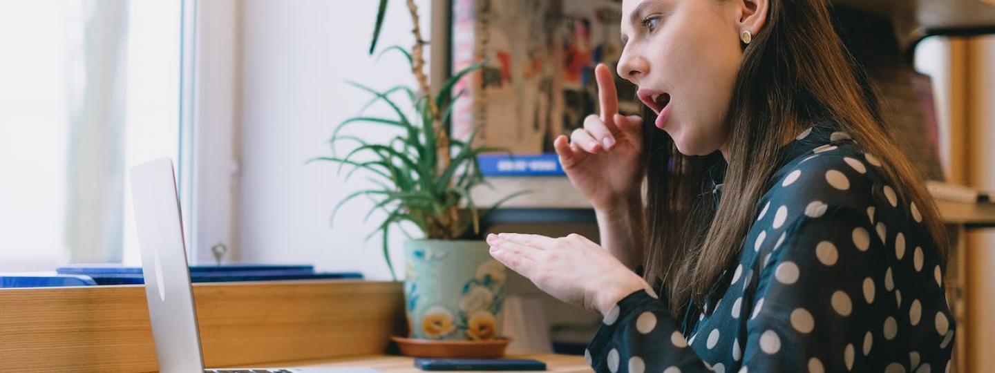 A woman with brown sitting by a window in front of a laptop, using sign language hand gestures. She is wearing a balck and white polka dot top, and a green plant sits on her desk