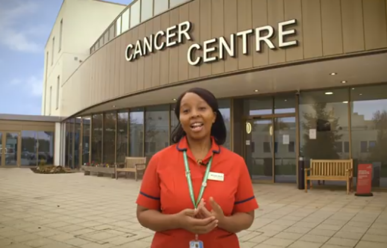A woman wearing an orange top standing outside a building with a sign above her that says 'Cancer Centre' 