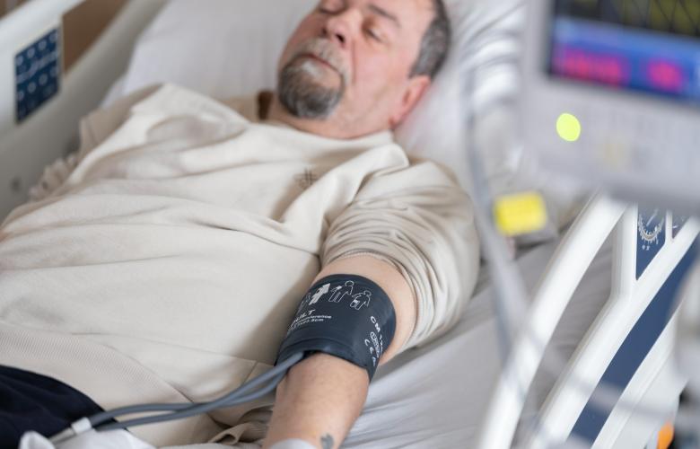 A man with a grey beard, wearing a beige t shirt lying in a hospital bed with his eyes closed. He has a blood pressure cuff on his arm.