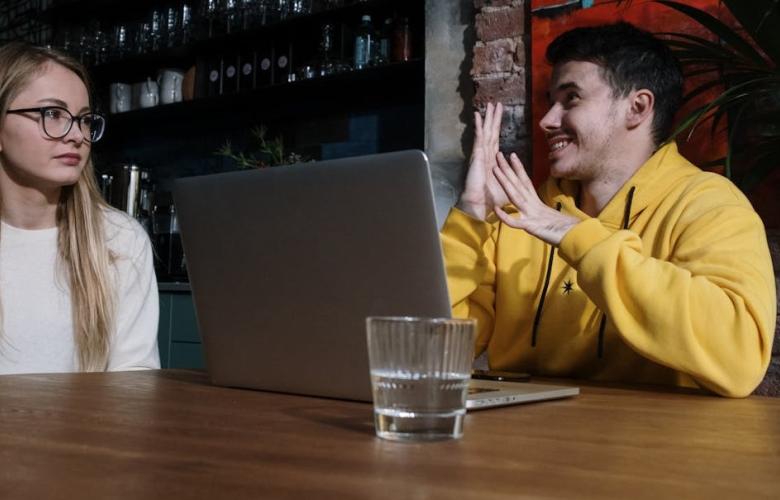 A woman on the left and a may on the right who is signing. He is wearing a yellow hoodie, sitting in front of a laptop.
