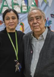 An Asian woman and older man stand side by side. She is wearing a black jumper with a lanyard around her neck, he is wearing a grey shirt and knitted waistcoat. The background behind them is bright blue with hand painted flowers.