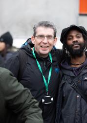 An older white man and younger black stand side by side outside smiling at the camera. They are wearing coats and name lanyards. It looks cold.  